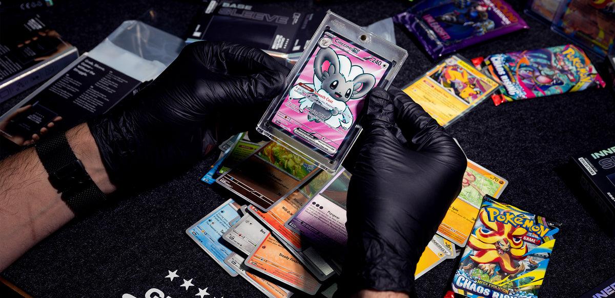 Two hands holding a Pokémon card in a magnetic sleeve. Several booster packs can be seen in the background.